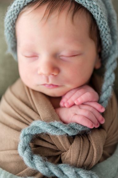 baby mit Haube gepuckt in naturtõnen Neugeborenes Baby, sanft in ein braunes Tuch gewickelt, mit einer blauen Mütze.