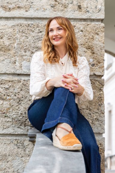 Business Brand fotografie Frau mit langen, braunen Haaren sitzt lächelnd auf einer Steinmauer. Trägt eine helle Bluse.