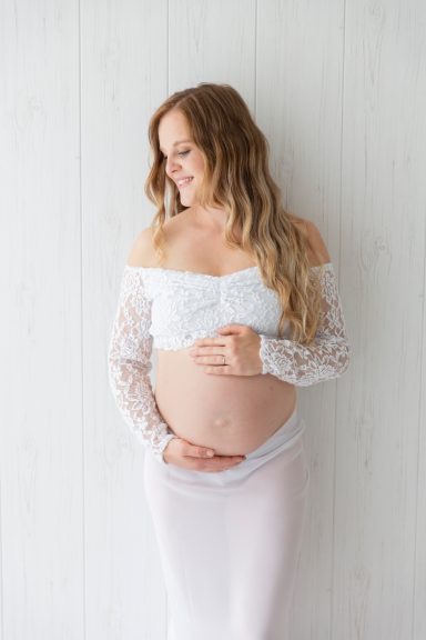 babybauchfotografie Schwangere Frau steht lächelnd vor einer hellen Wand und hält ihren Bauch.