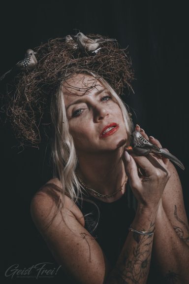 Vogelnest Frau mit blonden Haaren und Nest am Kopf hält einen kleinen Vogel in der Hand.